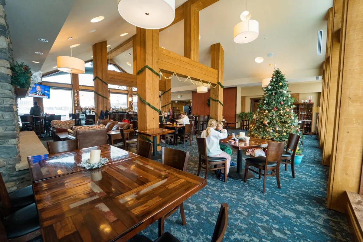 A festive gathering space with large wooden beams, a beautifully decorated Christmas tree, cozy dining areas, and a bar. The area feels warm, communal, and full of holiday spirit, suggesting a perfect venue for celebrations and family bonding.