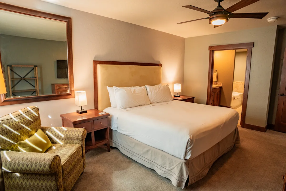 A serene bedroom scene with a cozy bed, inviting armchair, nightstands, and a glimpse into the bathroom. Natural light fills the space, suggesting comfort and relaxation after a day spent outdoors.