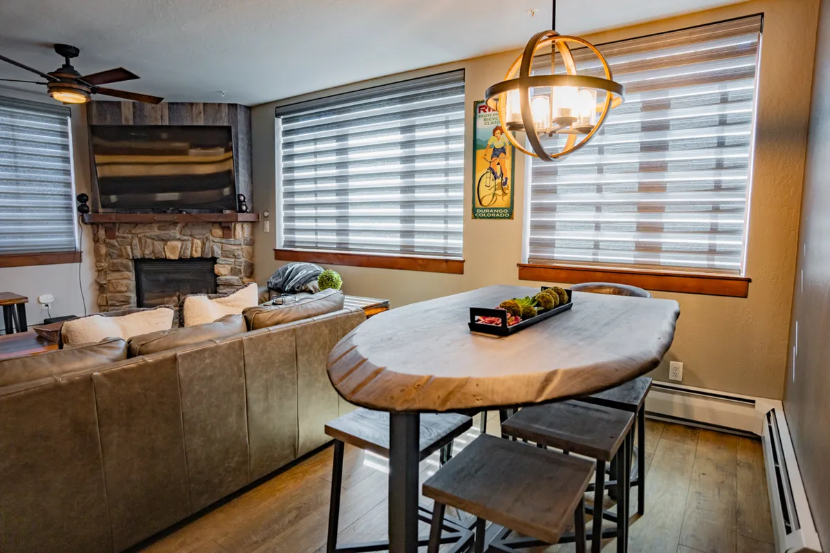A spacious living and dining area featuring a prominent stone gas fireplace, a rustic wooden dining table with ample seating, a comfortable couch, and decorative wall art. The scene radiates warmth and sociability, suggesting a perfect space for group gatherings and cozy evenings.