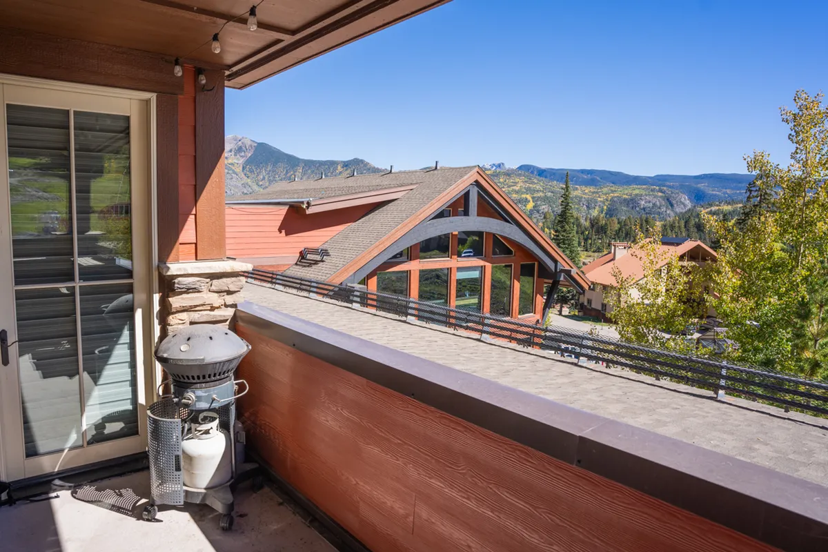 This exterior image showcases a balcony with a railing overlooking mountain views and neighboring houses, along with a grill for outdoor dining. It captures the opportunity to enjoy the fresh mountain air and scenic vistas right from your residence.