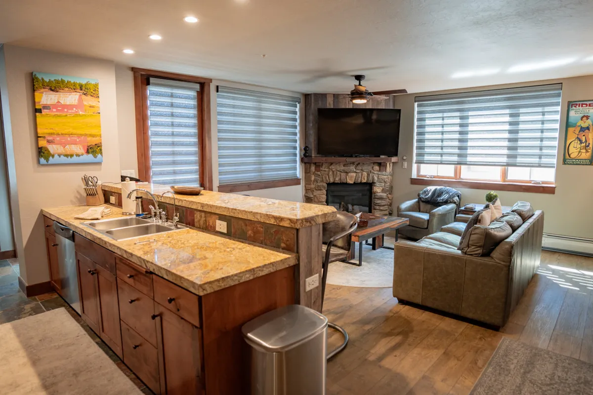 A view of a comfortable condo living space with a kitchen island, fireplace, and flexible seating arrangements. Artwork adds interest to the walls, and the layout is open, encouraging easy movement and conversation among guests.