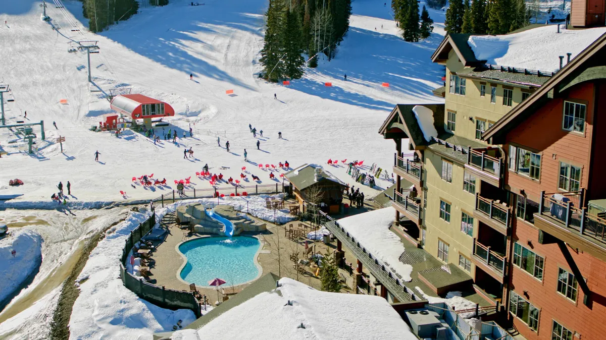 A vibrant exterior scene showcasing a resort with a heated swimming pool, skiers and snowboarders enjoying the slopes, and the main resort buildings. The pool is visible in the foreground, with snow-covered ski slopes and active guests creating a lively, energetic atmosphere.