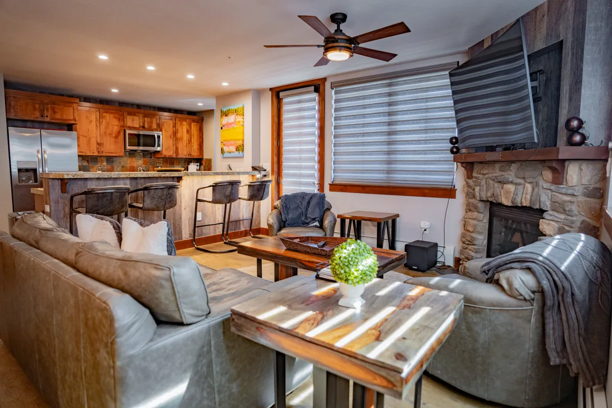 A spacious, open-plan living area featuring a comfortable sofa, a wooden coffee table, a modern kitchen island, and a cozy fireplace. Large windows in the background let in natural light and suggest scenic views just outside. The layout is ideal for group gatherings, relaxing after a day of adventure, or sharing drinks and stories around the fire.