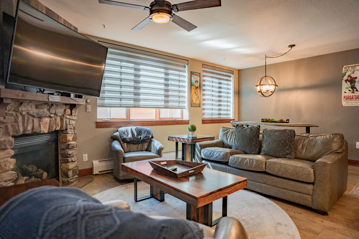 A warmly lit living area featuring a rustic stone fireplace, a comfortable seating arrangement, and a TV mounted above the mantel. Wooden accents and a large coffee table contribute to a cozy yet spacious feel, perfect for relaxing after a day exploring the mountains.