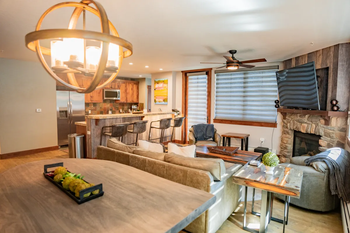 The image showcases a spacious and elegantly furnished open-concept living area inside a luxury condo. A sparkling chandelier hangs over a cozy seating area with a plush sofa, while a stone fireplace glows nearby. The modern kitchen features granite countertops, a large island with bar stools, and seamlessly connects to the dining area—creating a perfect setting for gatherings, celebrations, or relaxing after a day on the slopes.