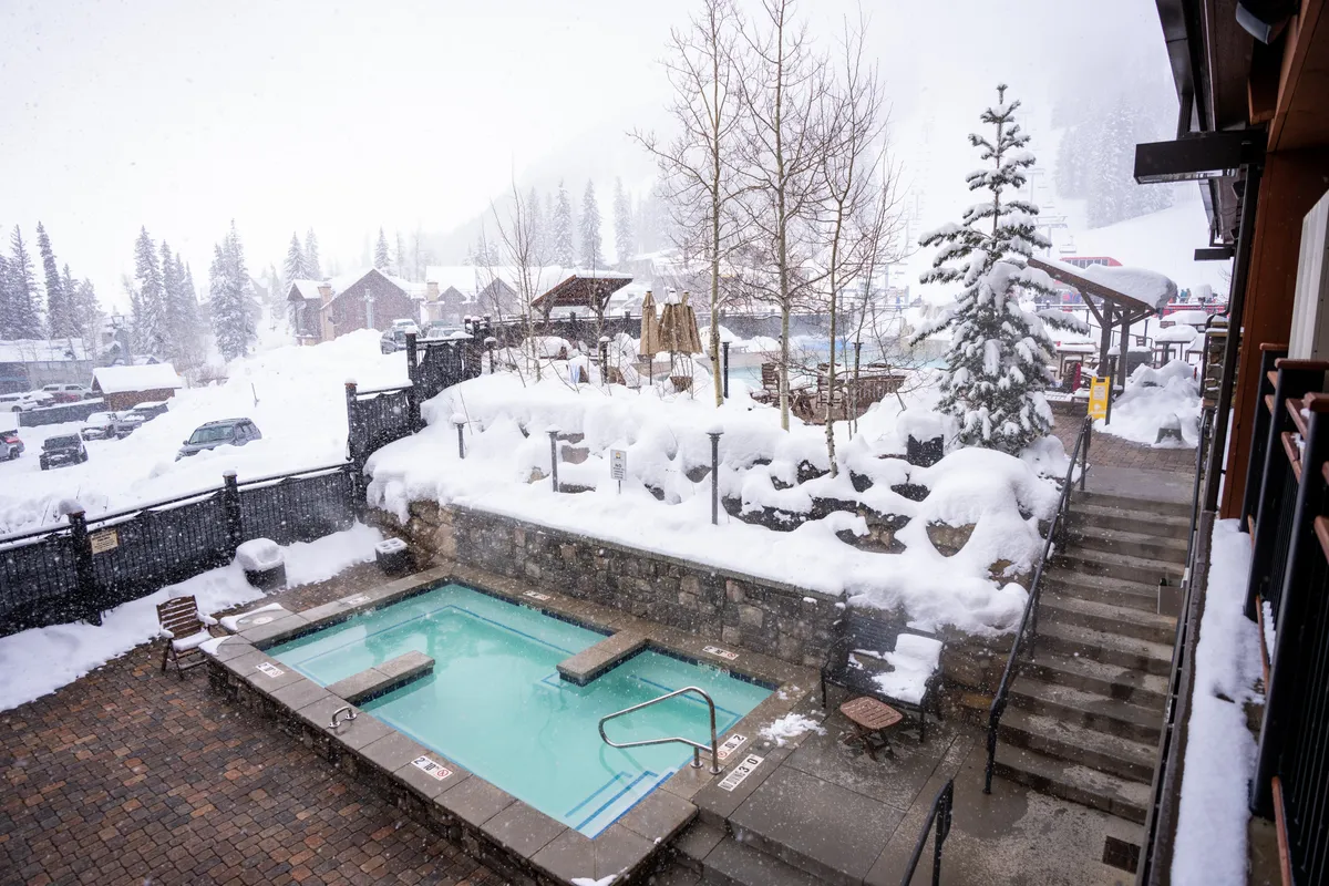 This exterior image features a steaming hot tub surrounded by a snow-covered landscape and tall trees. Steps lead up to the stone-walled hot tub area, inviting relaxation after a day on the slopes. The snowy setting and outdoor spa evoke the tranquil après-ski experience.