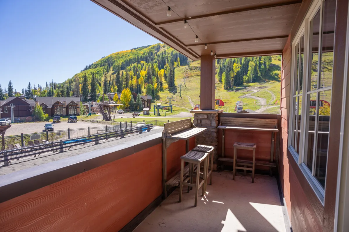 An exterior balcony setup with comfortable seating and a view of the surrounding mountains, trees, and a ski lift. The mood is relaxed, inviting guests to enjoy sunrise coffee or evening gatherings with a spectacular backdrop.