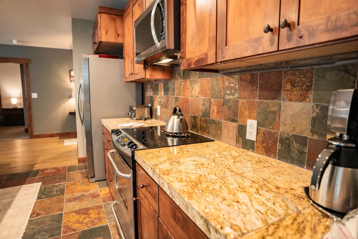 This image features a kitchen with a prominent granite countertop, warm wooden cabinetry, a stylish slate backsplash, and modern stainless steel appliances. Pots, pans, and utensils are visible, emphasizing the kitchen's readiness for serious cooking and gatherings.