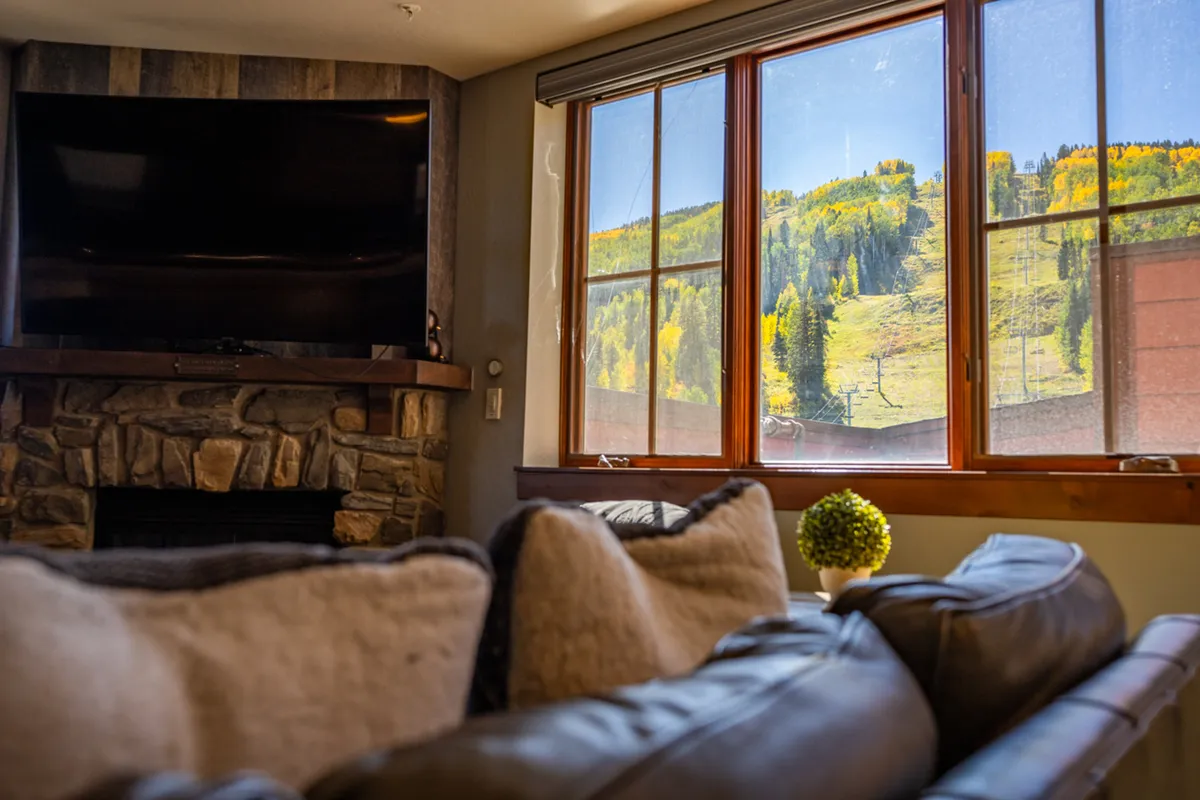 A living room with a large window framing stunning mountain views, a plush couch adorned with pillows, a stone fireplace, and a television. The setting is intimate and inviting, inviting guests to relax while taking in the Needles Mountain Range.