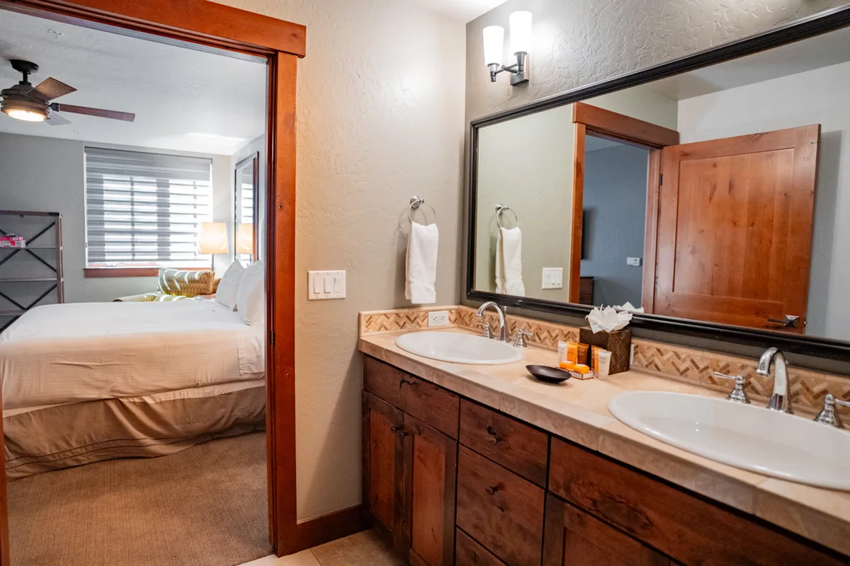 A spa-inspired bathroom with a double sink vanity, expansive mirror, warm wooden cabinetry, and a partial view into the adjoining bedroom. The atmosphere is calm and rejuvenating, perfect for morning or evening routines.
