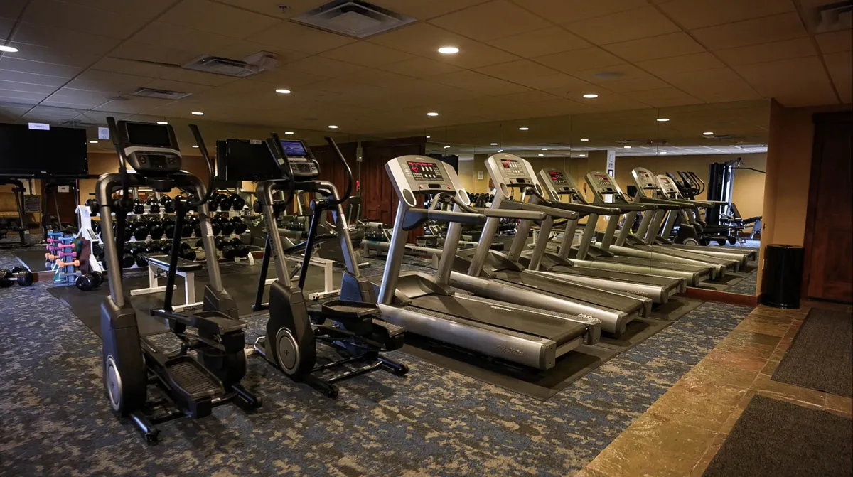 A modern interior fitness center with elliptical machines, treadmills, dumbbells, and mirrored walls. The space is bright, energetic, and designed for both individual workouts and group fitness sessions.